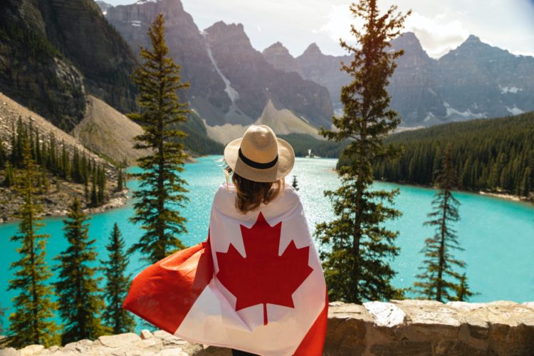 A woman looks out over a lake, mountains and trees, her back is to the camera, and she wears a hat. She is wrapped in a Canadian flag.