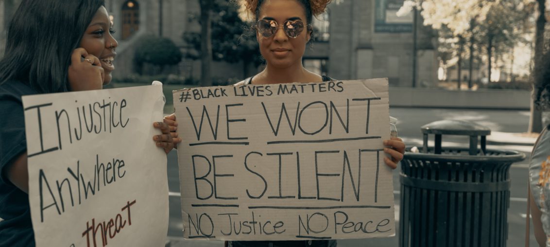 Two smiling black women stand holding handmade signs reading #Blacklives matter We Won't Be Silent NO Justice NO Peace and Injustice Anywhere is a Treat to Justice Everywhere