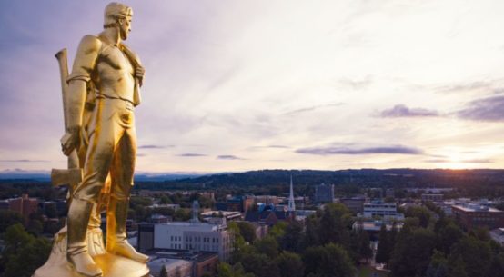 A golden statue of a male dressed as a pioneer holding an ax is seen atop the capitol in Salem Oregon, high above the tree lined mountain horizon