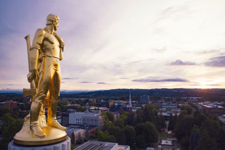 A golden statue of a male dressed as a pioneer holding an ax is seen atop the capitol in Salem Oregon, high above the tree lined mountain horizon
