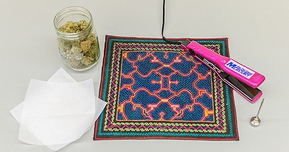 A jar of cannabis buds sits next to a hair straightening iron and some parchment paper