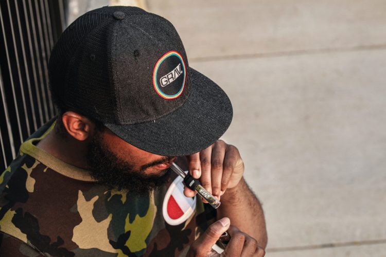 A black man in a baseball cap holds a lighter up to a glass one hitter