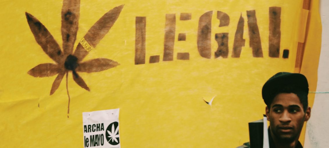A young Black man with a backpack stands in front of a yellow banner behind him which reads Legal next to a cannabis leaf