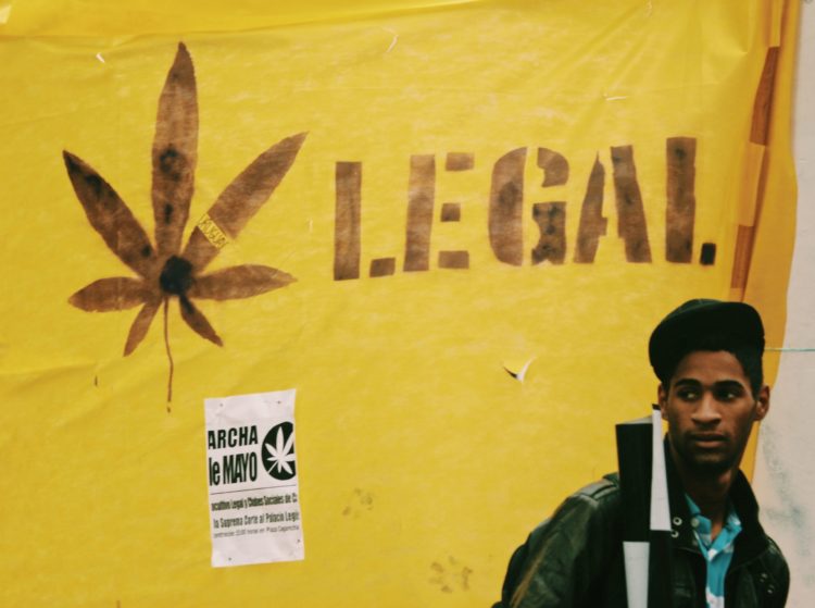 A young Black man with a backpack stands in front of a yellow banner behind him which reads Legal next to a cannabis leaf