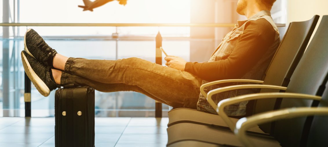 A man sits in a row of chairs at an airport, his feet propped up on a piece of luggage. He looks out a window at a plane taking off.