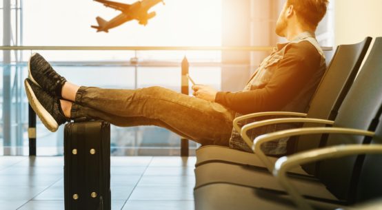 A man sits in a row of chairs at an airport, his feet propped up on a piece of luggage. He looks out a window at a plane taking off.