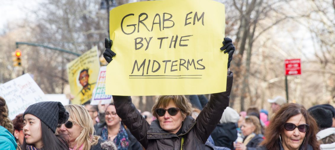 I middle aged woman wearing a black jacket and dark sunglasses holds up a yellow handmade sign reading Grab Em By The Mid Terms. she is in the middle of the Million Woman March.