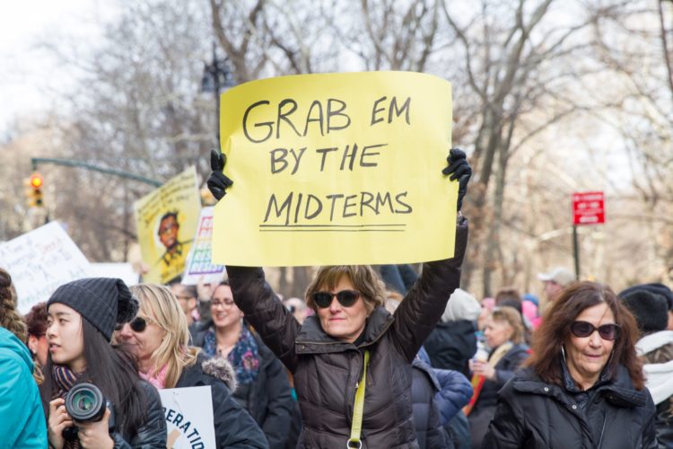 I middle aged woman wearing a black jacket and dark sunglasses holds up a yellow handmade sign reading Grab Em By The Mid Terms. she is in the middle of the Million Woman March.