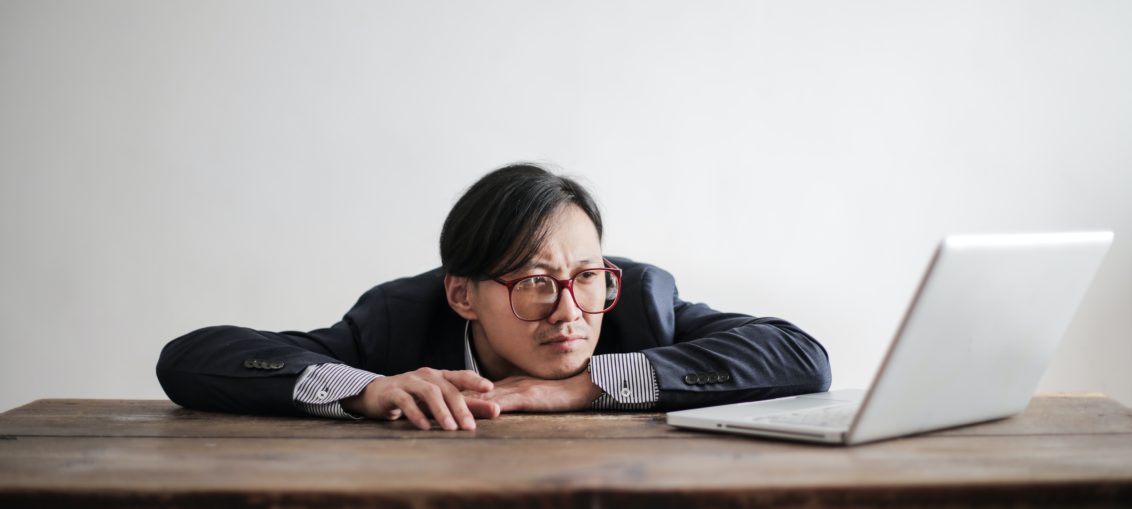 A bored man sits at a desk looking at a laptop