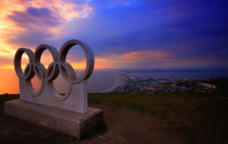 A statue of the five Olympic Rings sits above the ocean at sunset