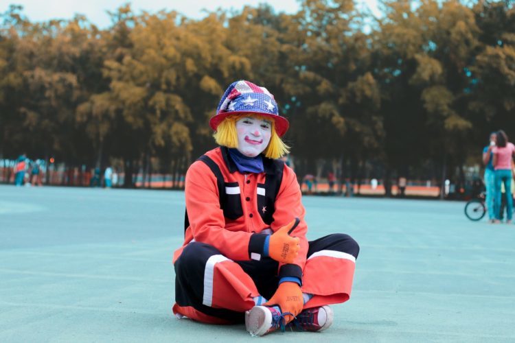 A sitting clown gives the thumbs up sign wearing a US flag hat